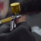 A close-up of an airbrush paint gun with a gold cap and metallic body, held by a hand wearing a black glove. The background is heavily blurred, showing what appears to be a person's head and hair.